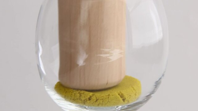 Kinetic sand ASMR compression. Wooden pestle presses yellow sand in glass bowl, creating circular indentation patterns as sand compresses.