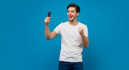 Fototapeta premium Excited young man celebrating success while using smartphone against vibrant blue background