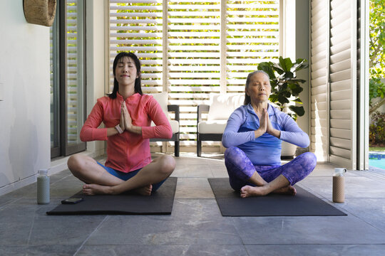 Meditating Asian mother and daughter sitting on yoga mats on terrace, with water bottles - Powered by Adobe