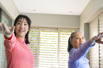 Stretching Asian female workout partners performing forward arm stretch in home gym, with blinds