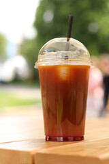 Bumble coffee in disposable cup on wooden table, close-up. Iced coffee outdoors with blurred park in background. Cold coffee in plastic disposable cup with straw. Takeaway drink
