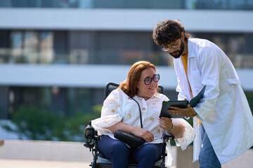 Doctor showing tablet to smiling woman in wheelchair outdoors - wheelchair and disability concept