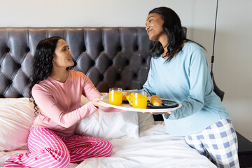 Sharing breakfast Diverse female friends sitting in modern bedroom, passing tray with croissants