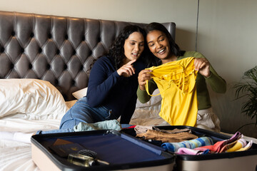 Diverse female friends holding bright yellow top while sitting on bed, with open suitcase