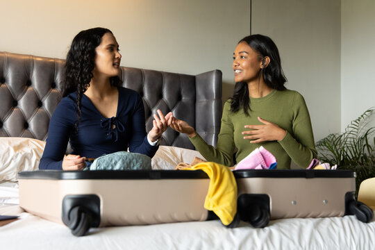 Diverse female friends packing hard-shell suitcase in bedroom, with assorted clothing items