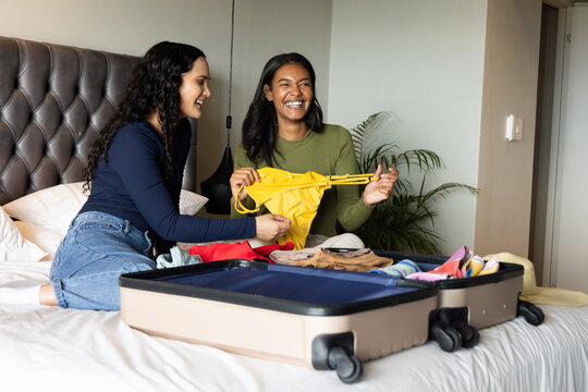 Diverse female friends sorting bikini top and clothes on bed in bedroom, with rolling suitcase - Powered by Adobe