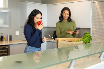 Diverse female friends smelling pepper and unpacking groceries at kitchen island with phone