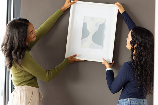 Positioning Diverse female friends holding white-framed abstract painting on taupe wall at home