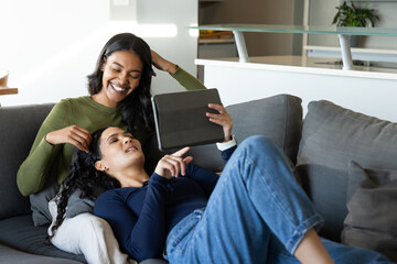 Relaxing two women sharing grey sofa in bright living area, with tablet and throw pillows