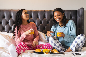 Laughing Diverse female friends sitting cross-legged on bed, with breakfast trays and orange juice