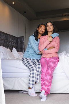 Hugging diverse female partners sitting on bed in modern bedroom, with floor lamp glow