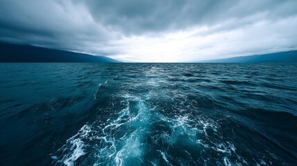 Fototapeta premium Serene Ocean View Under Dark Clouds - A tranquil ocean scene under a dramatic, dark sky. The water is deep blue, with a wake trailing behind