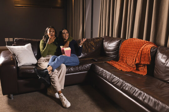 Watching diverse female friends sitting on sofa at home, with striped popcorn tub, copy space