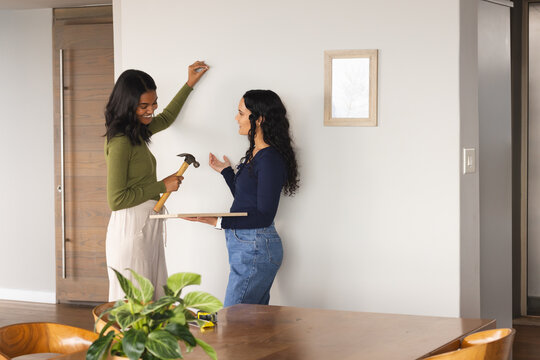 Hanging frame Diverse female friends installing wall art at home, with hammer and tape measure