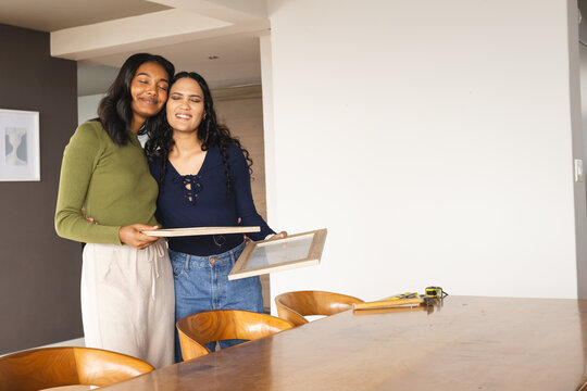Hugging diverse female friends holding empty wooden frames at modern dining table, copy space