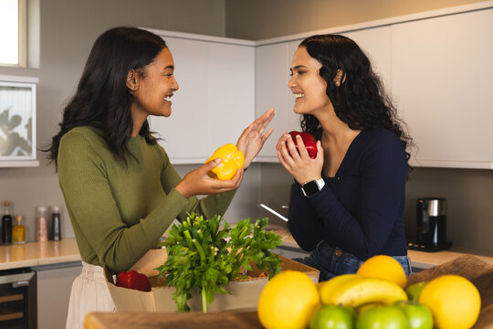 Smiling Diverse female friends unpacking groceries at home kitchen island, with peppers and herbs