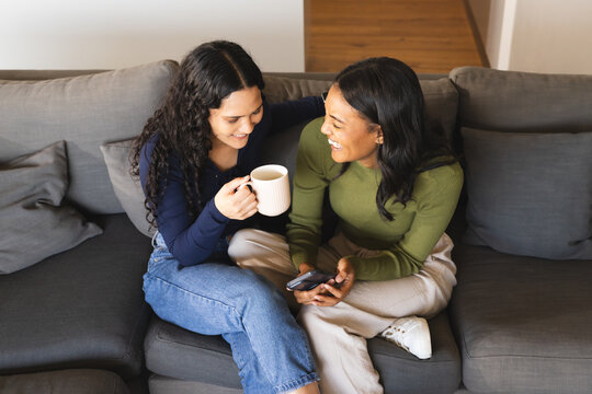 Laughing diverse female friends sitting on gray sofa in living room, holding mug and smartphone