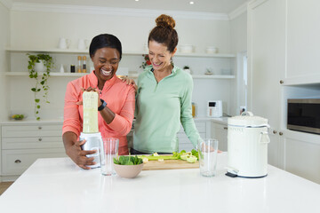 Blending green smoothie by diverse female friends at kitchen island, with blender container