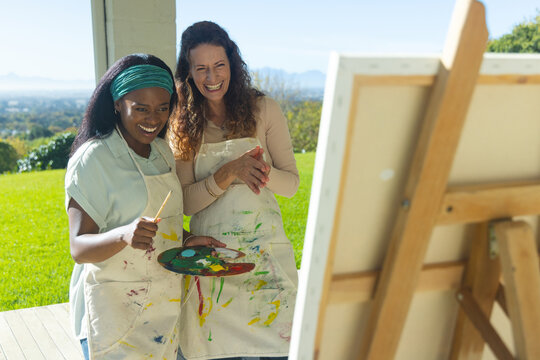 Diverse female friends blending colors on wooden patio, with easel and palette, copy space