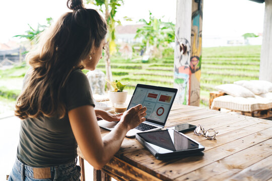 Young brunette freelancer checking stats on laptop in outdoor coworking space with lush green rice field view, multitasking with tablet, eco-conscious modern digital workspace setup
