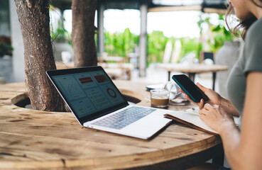 Freelancer working on laptop and taking notes in notebook while holding smartphone in outdoor café, surrounded by coffee, headphones, and greenery, blending tech and comfort