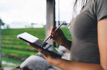 Close-up of woman’s hands drawing on digital tablet with stylus in tropical coworking zone,...