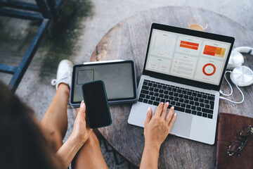 Overhead shot of freelancer holding smartphone and working on laptop with tablet nearby, surrounded by gadgets, headphones, and coffee in modern digital workspace