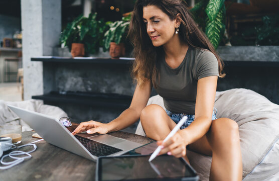 Focused female freelancer working with laptop and tablet simultaneously, analyzing charts with stylus in cozy indoor workspace surrounded by plants