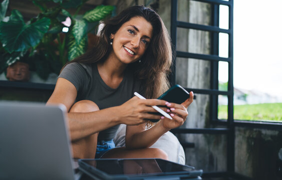 Attractive young woman smiling and looking at camera while holding phone and stylus, sitting relaxed in modern workspace with tablet and laptop
