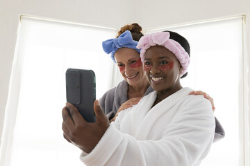 Smiling diverse female friends taking selfie in spa room, with smartphone and under-eye patches