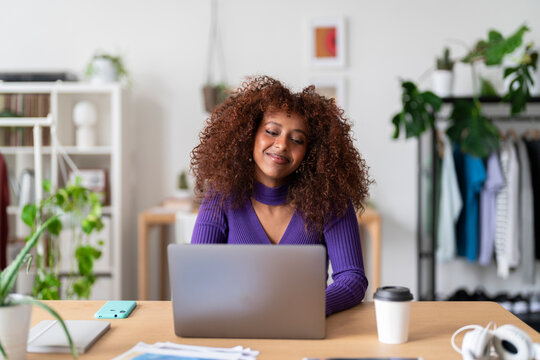 A young woman with curly hair is working on her laptop at a desk in a bright room. She appears focused and content, likely engaged in remote work or online learning from the comfort of her home.