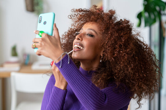 A woman with curly hair applies lip gloss while looking at her phone. She is likely checking her makeup or recording a beauty tutorial video.
