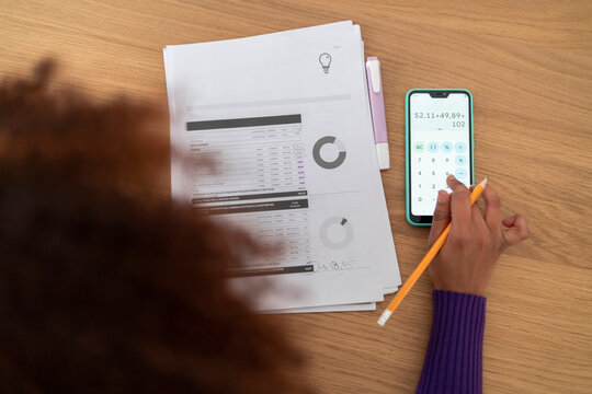 A person uses a phone calculator to check figures on financial documents. They are likely managing expenses or verifying calculations for personal or business purposes.