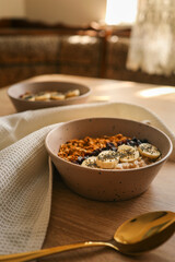 Top view of ceramic bowl filled with oatmeal, sliced bananas, dried cherries, granola and chia seeds, placed on a wooden surface with a spoon and kitchen towel