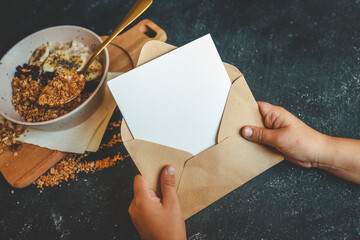 Hands holding kraft envelope with blank card next to bowl of granola and banana on dark rustic background. Cozy breakfast moment with handwritten note or greeting