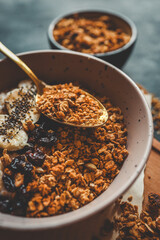 Top view of ceramic bowl filled with oatmeal, sliced bananas, dried cherries, granola and chia seeds, placed on a wooden surface with a spoon and kitchen towel