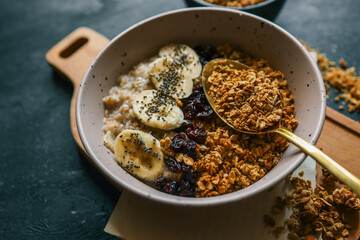 Top view of ceramic bowl filled with oatmeal, sliced bananas, dried cherries, granola and chia seeds, placed on a wooden surface with a spoon and kitchen towel