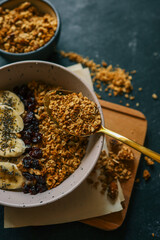 Child’s hand holding a spoon with granola over a bowl of oatmeal with banana slices and dried berries on a dark background. Healthy breakfast concept with dramatic lighting