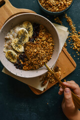 Child’s hand holding a spoon with granola over a bowl of oatmeal with banana slices and dried berries on a dark background. Healthy breakfast concept with dramatic lighting