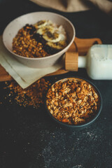 Top view of oatmeal bowl with banana, chia seeds, dried cherries, and granola, placed on wooden board with spilled granola and milk bottle on dark background
