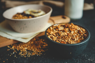 Granola in black ceramic bowl placed on dark surface with oatmeal bowl, milk bottle, and wooden board in soft background, rustic natural morning setting
