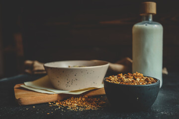 Granola in black ceramic bowl placed on dark surface with oatmeal bowl, milk bottle, and wooden board in soft background, rustic natural morning setting