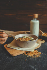 Granola in black ceramic bowl placed on dark surface with oatmeal bowl, milk bottle, and wooden board in soft background, rustic natural morning setting