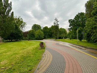 A curved paved pathway with benches in a peaceful urban park on a cloudy day. The path is...