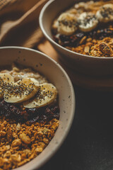 Flat lay of two ceramic bowls with creamy oatmeal, topped with granola, banana slices, dried berries, and chia seeds, placed on a wooden surface with beige waffle towel