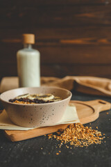 Granola in black ceramic bowl placed on dark surface with oatmeal bowl, milk bottle, and wooden...