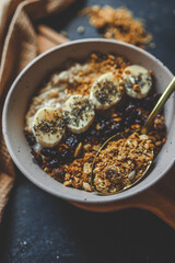 Top view of ceramic bowl filled with oatmeal, sliced bananas, dried cherries, granola and chia seeds, placed on a wooden surface with a spoon and kitchen towel