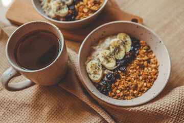 Top view of two ceramic bowls filled with oatmeal, banana slices, granola, and chia seeds, with a cup of tea and kitchen towel on dark textured table