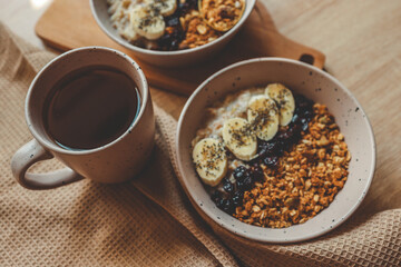 Top view of two ceramic bowls filled with oatmeal, banana slices, granola, and chia seeds, with a cup of tea and kitchen towel on dark textured table
