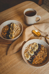 Warm morning setup with two bowls of oatmeal topped with banana, chia, dried berries, and granola, served with tea and golden spoon on wooden table with waffle cloth